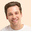 Young man with brown hair smiling at camera, wearing light-colored t-shirt against neutral background.