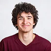 Portrait of a person with curly dark hair wearing a burgundy v-neck shirt against a white background.