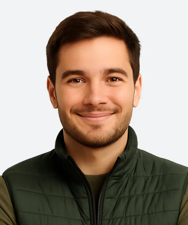 Man with dark hair and beard wearing a dark green quilted vest over a tan shirt, smiling at camera against white background.
