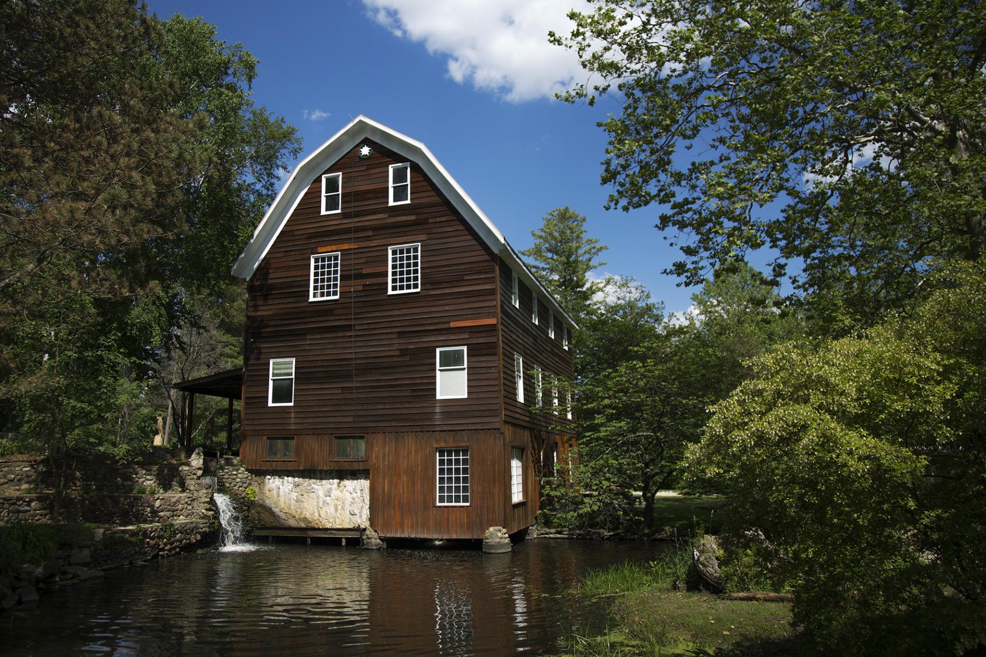 Historic brown mill building with white trim situated over a stream, surrounded by green trees under a blue sky.