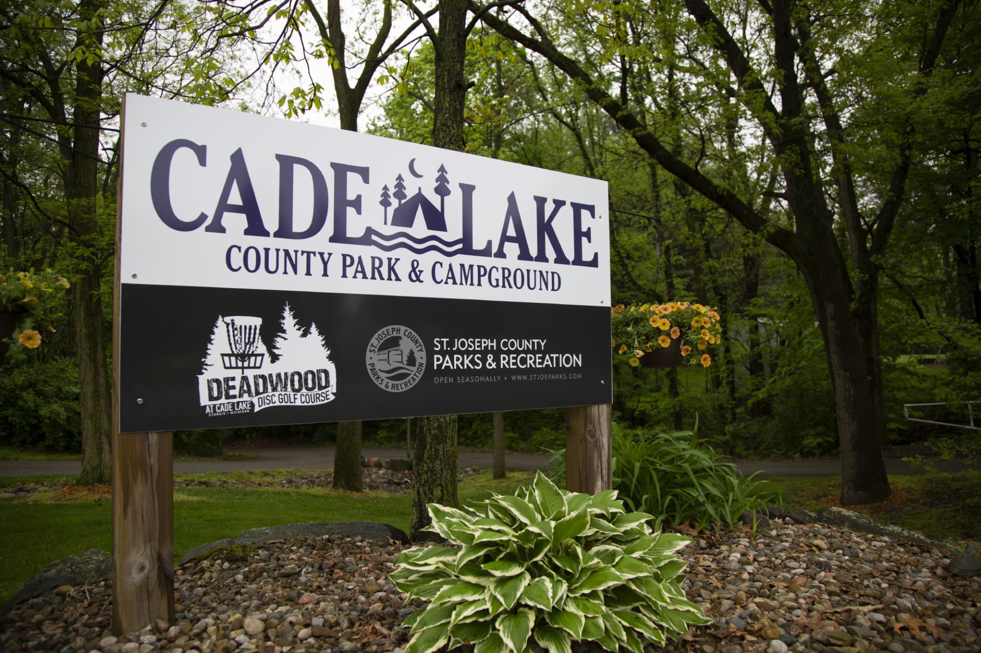 Cade Lake County Park & Campground entrance sign with Deadwood Disc Golf Course logo, surrounded by trees and landscaping.