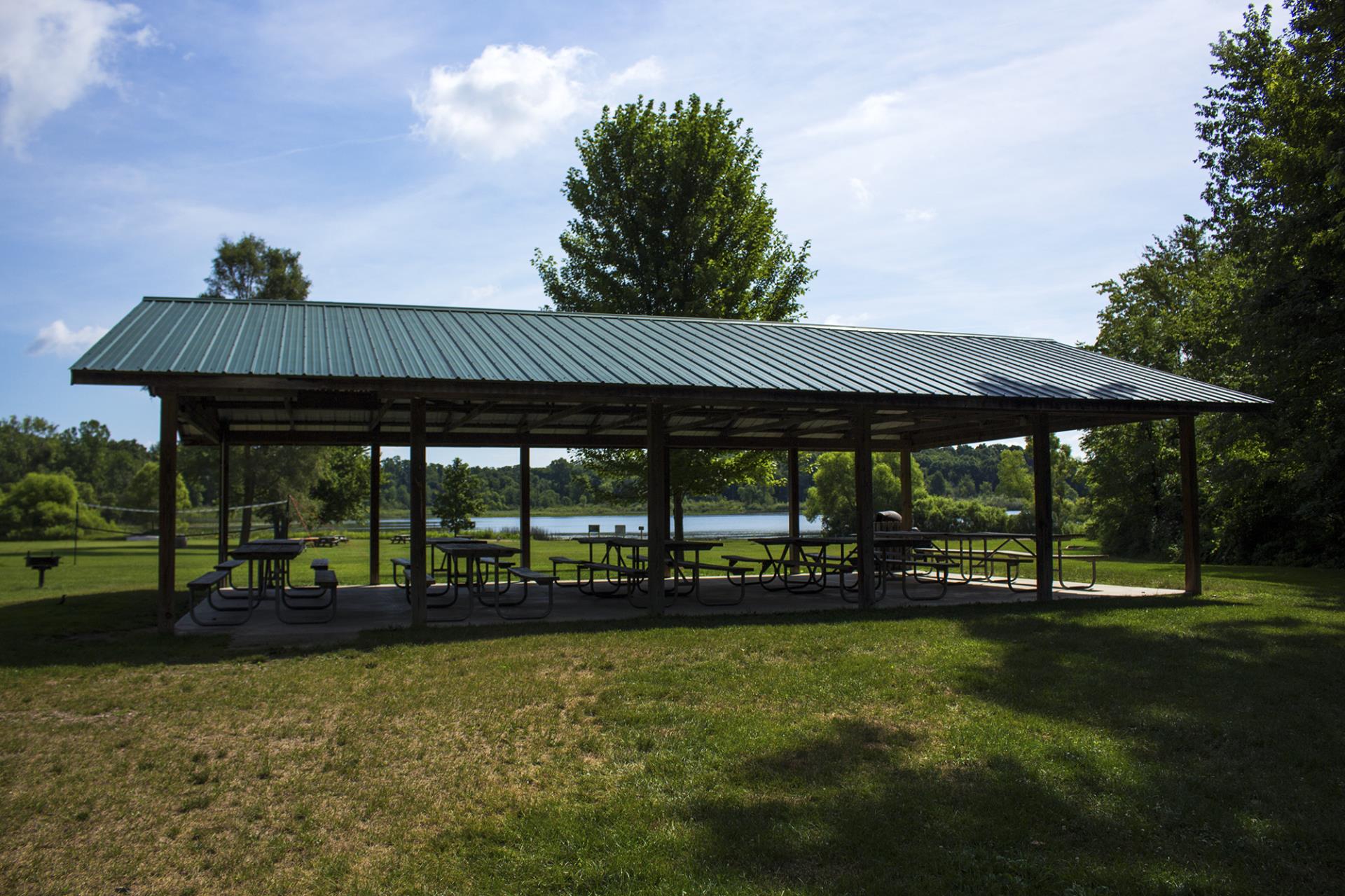 Large covered picnic pavilion with metal roof and picnic tables beside a river, surrounded by green grass and trees.