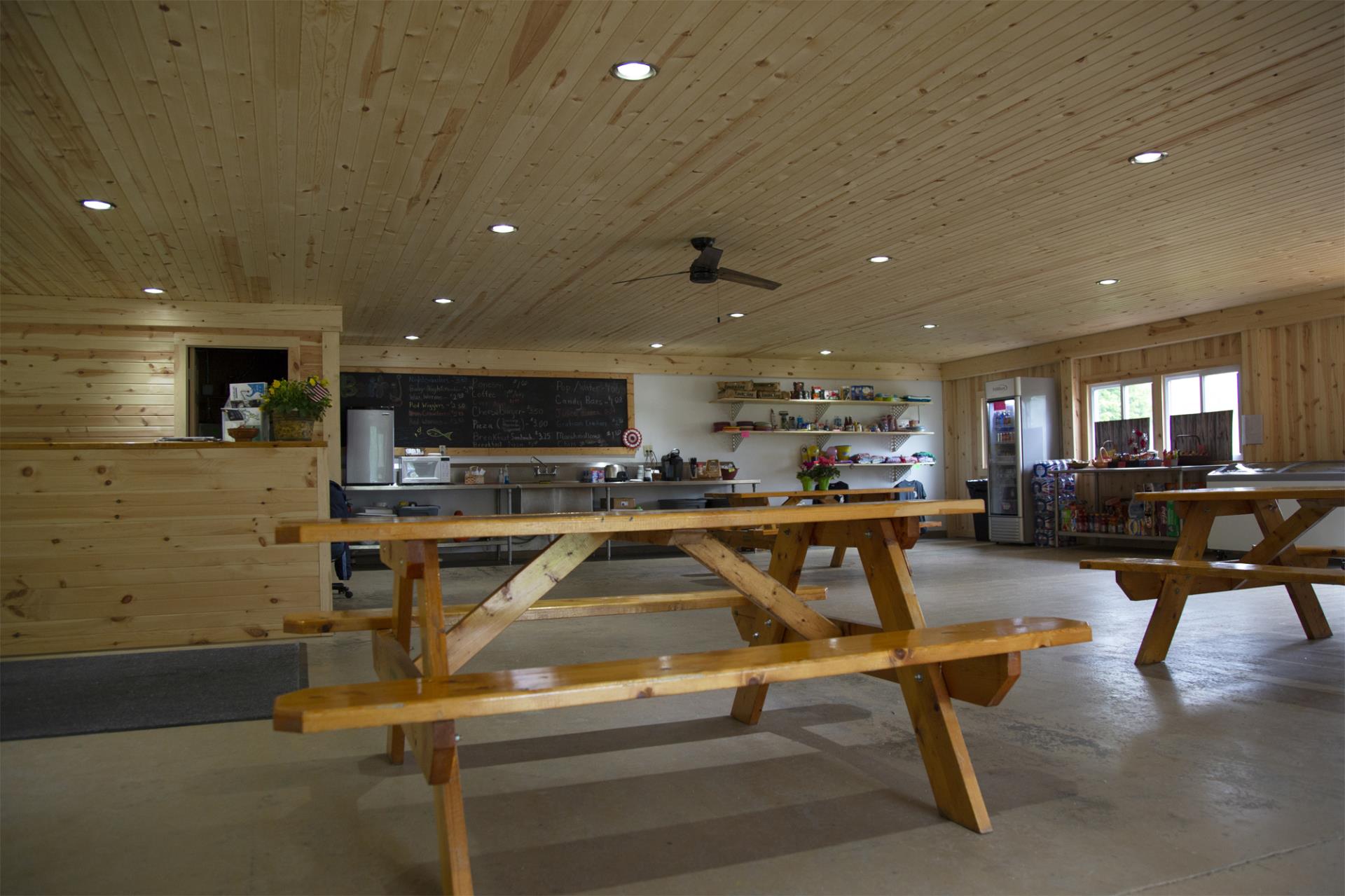 Large wooden building interior with picnic tables, kitchen area, and wood ceiling with recessed lighting.