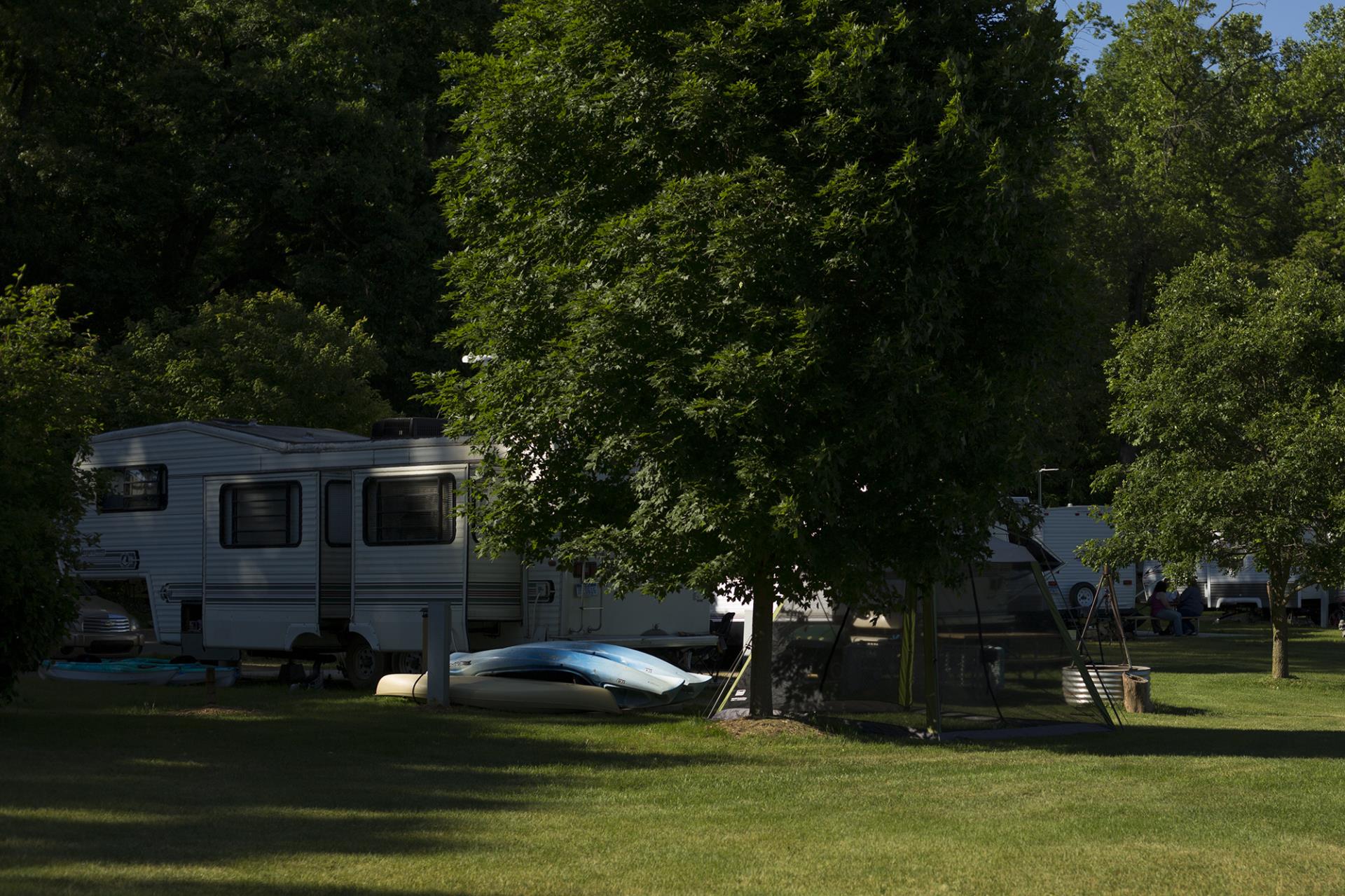 RV camper parked at campground with kayak underneath tree and picnic table nearby.