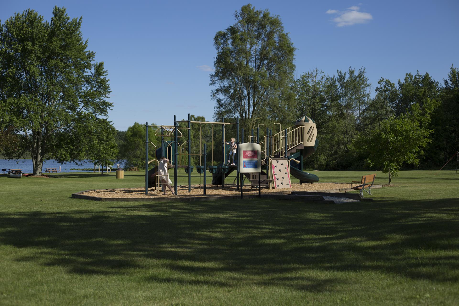 Playground with slide, climbing structures, and activity panels in a lakeside park with green grass and trees.