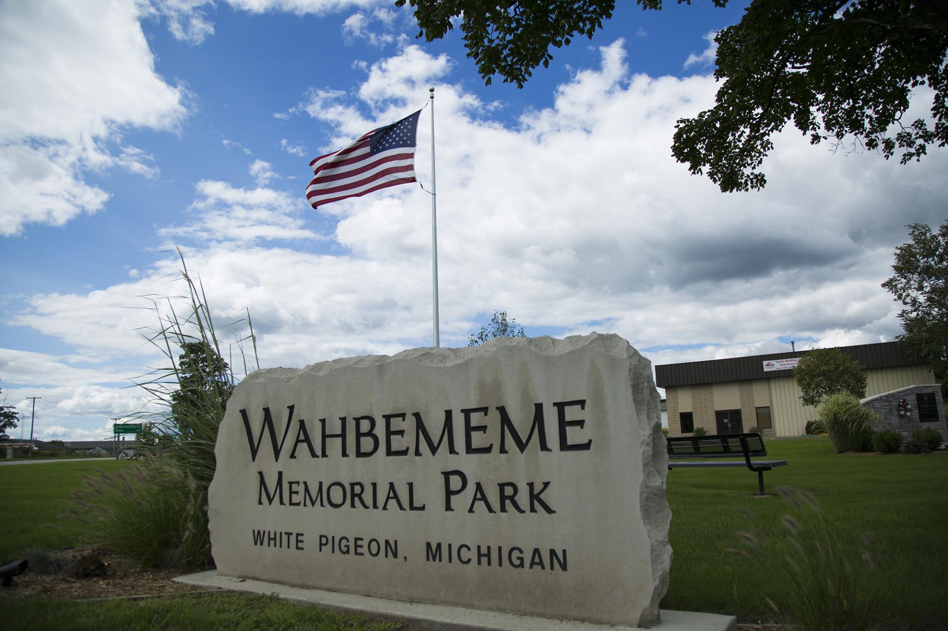 Wahbememe Memorial Park sign in White Pigeon, Michigan with American flag and building in background.