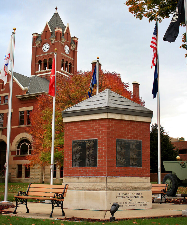 St Joseph County Veterans Memorial with brick structure, plaques, flags, and historic red courthouse tower in autumn.