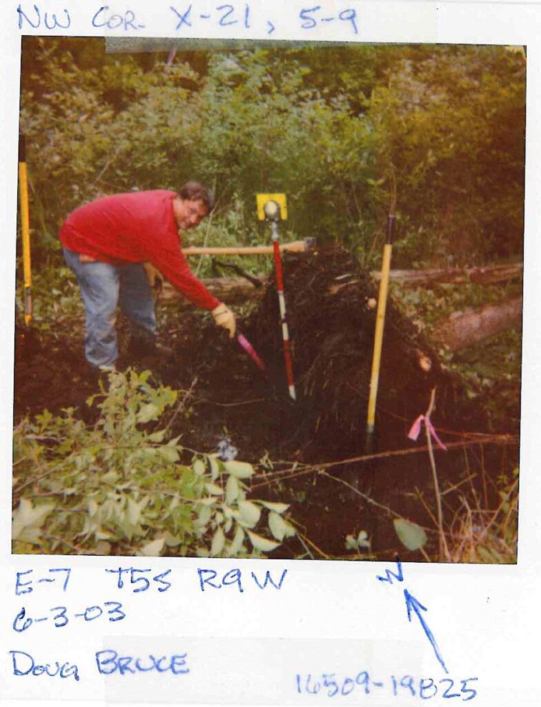 Man in red shirt conducting archaeological excavation with measuring tools and stakes in wooded area.