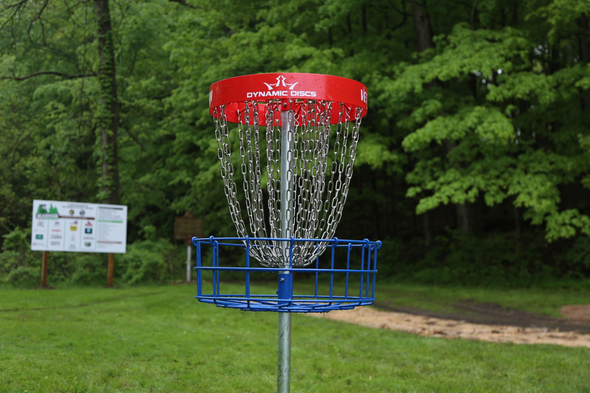 Dynamic Discs disc golf basket with red top and blue base in a wooded park setting with informational sign.