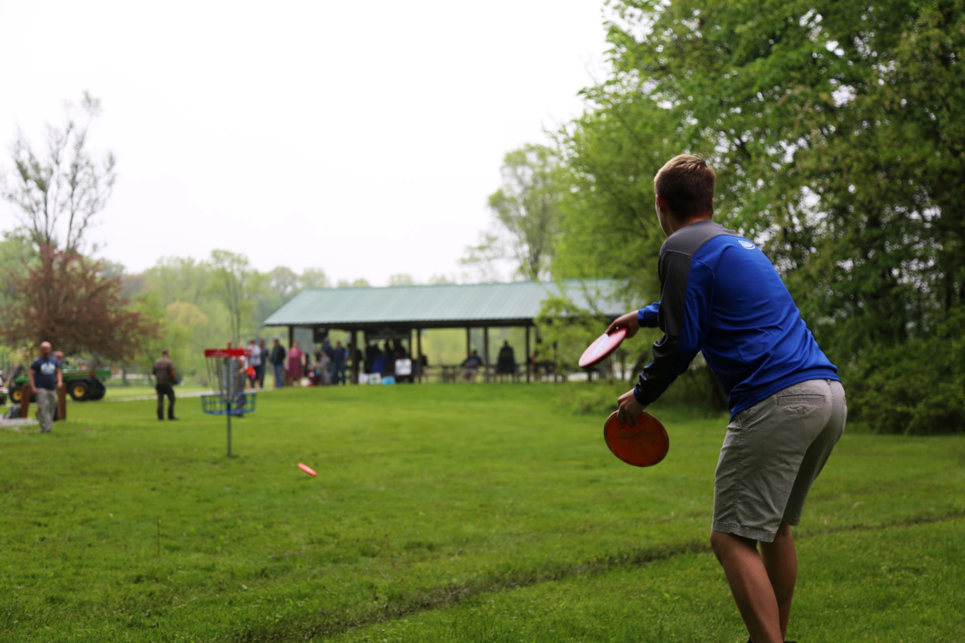 Man in blue shirt playing disc golf in a park with shelter and spectators in the background.