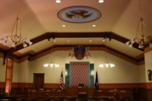 Courtroom interior with judge's bench, American flag, wooden furnishings, and ornate ceiling details.