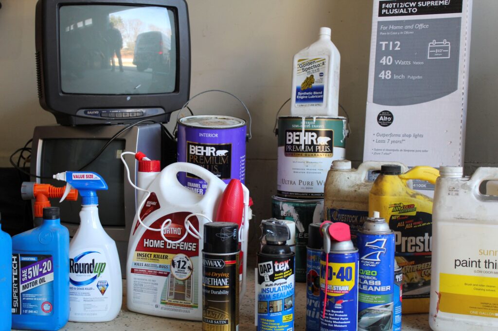 Cluttered shelf with cleaning supplies, paint cans, motor oil, and a vintage CRT television displaying an outdoor scene.
