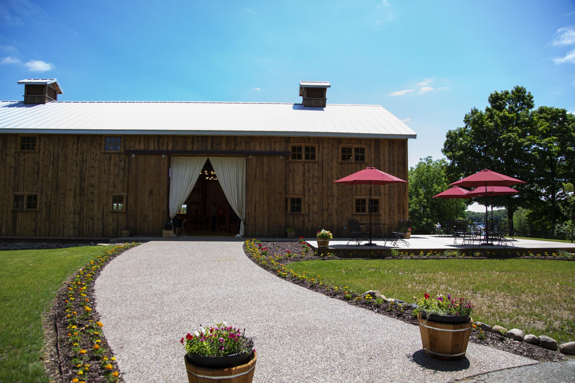 Rustic wooden barn with metal roof, white curtains, gravel driveway, landscaped grounds, and red umbrellas on patio area.