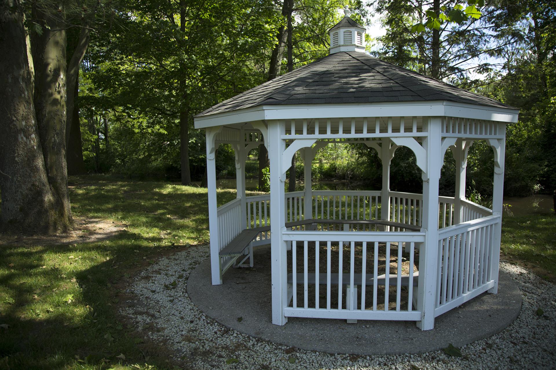 White octagonal gazebo with railed deck set on gravel in a wooded area surrounded by large trees.