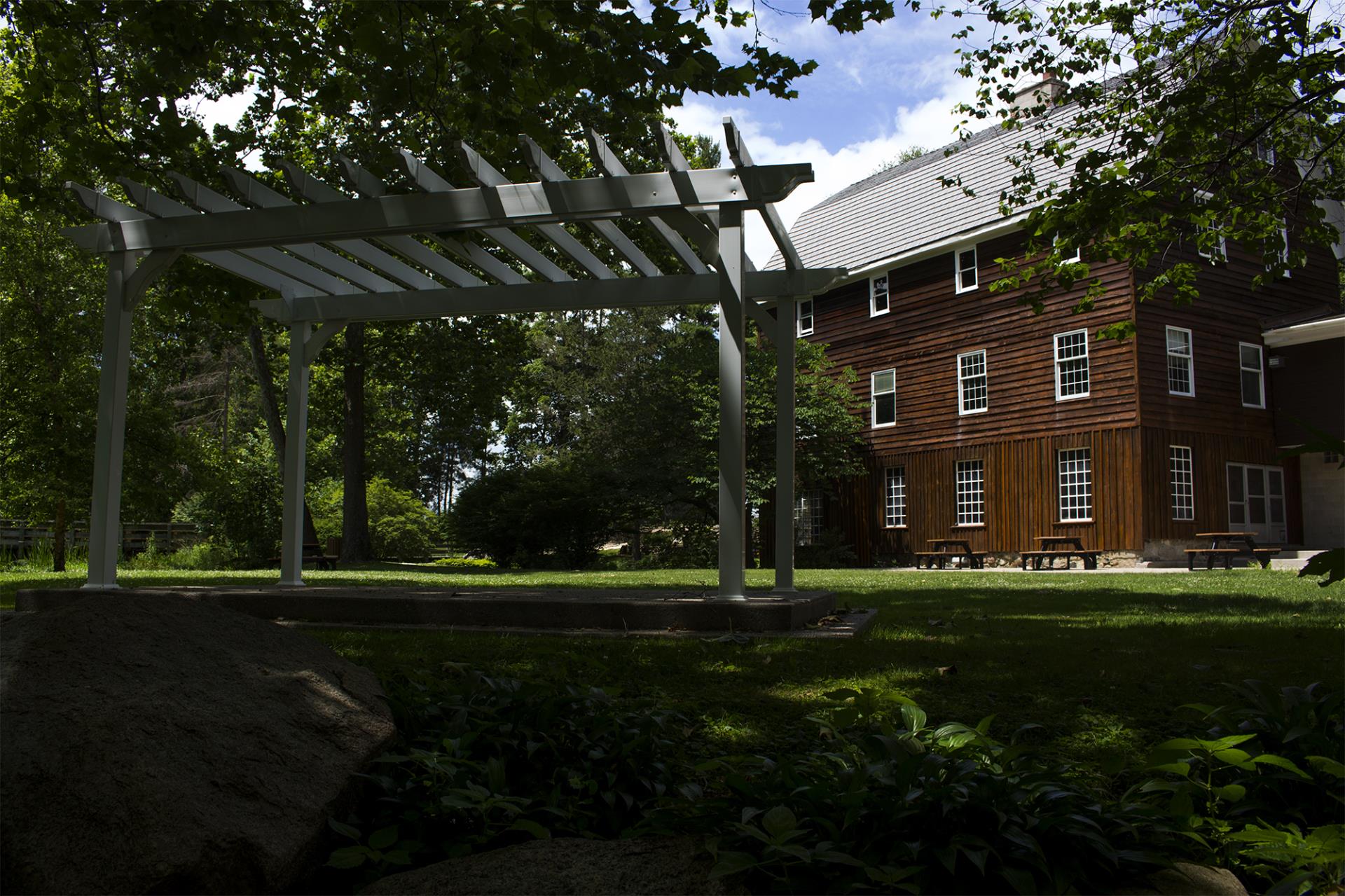 Modern gray pergola structure in front of a dark wooden barn with metal roof, surrounded by green trees and landscaping.