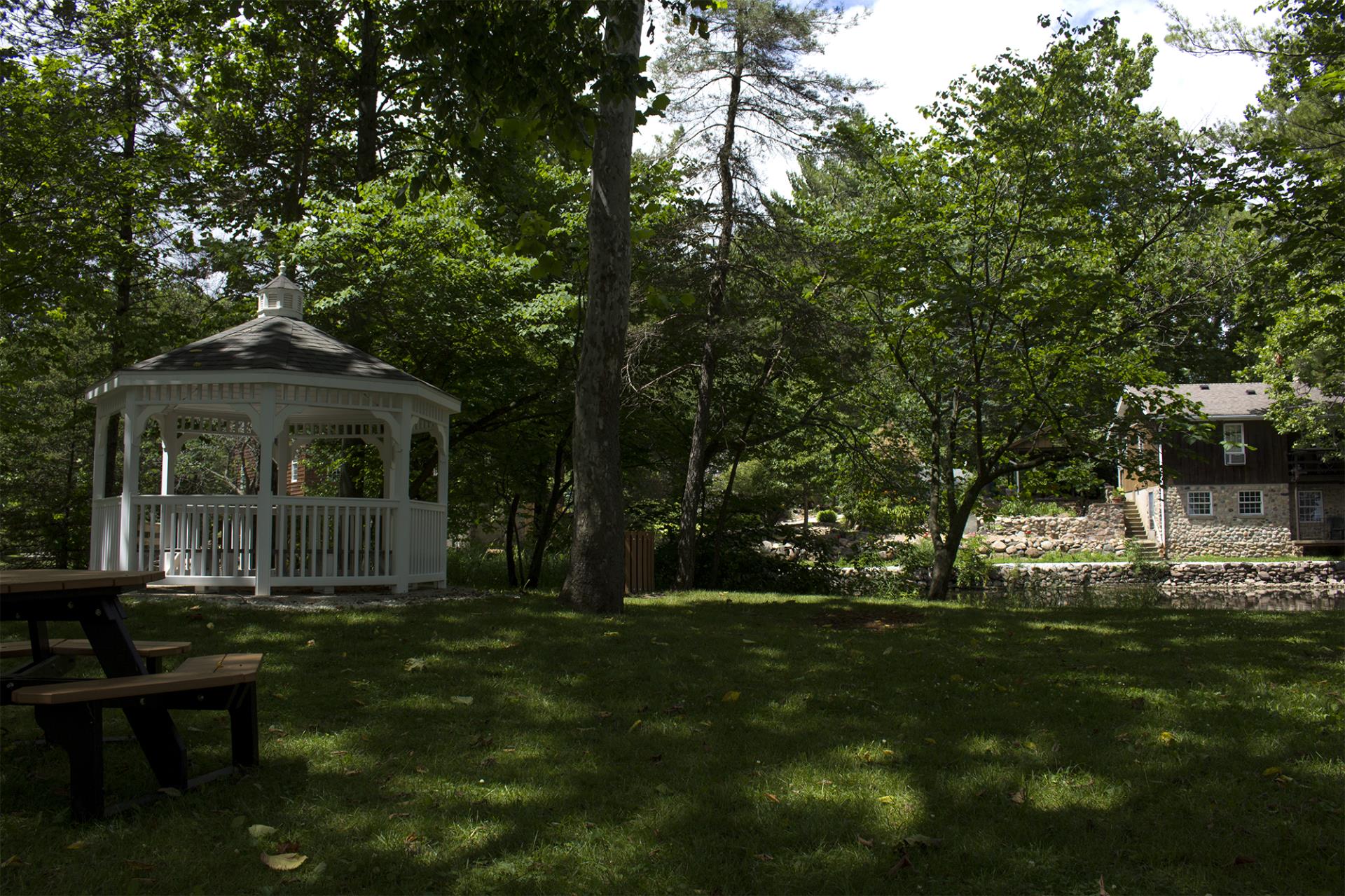 A white gazebo with black roof stands in a wooded park setting near a stone house and picnic table.