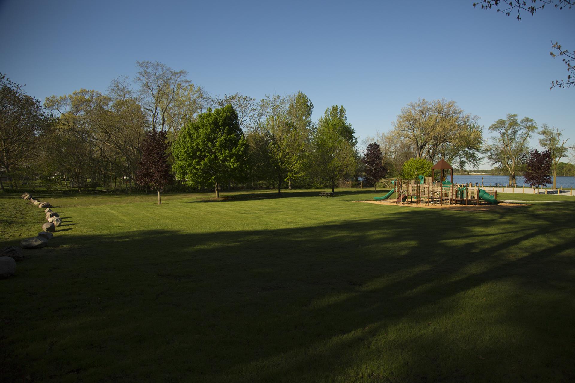 Spacious park with playground equipment, green lawn, mature trees, and water visible in background under clear sky.