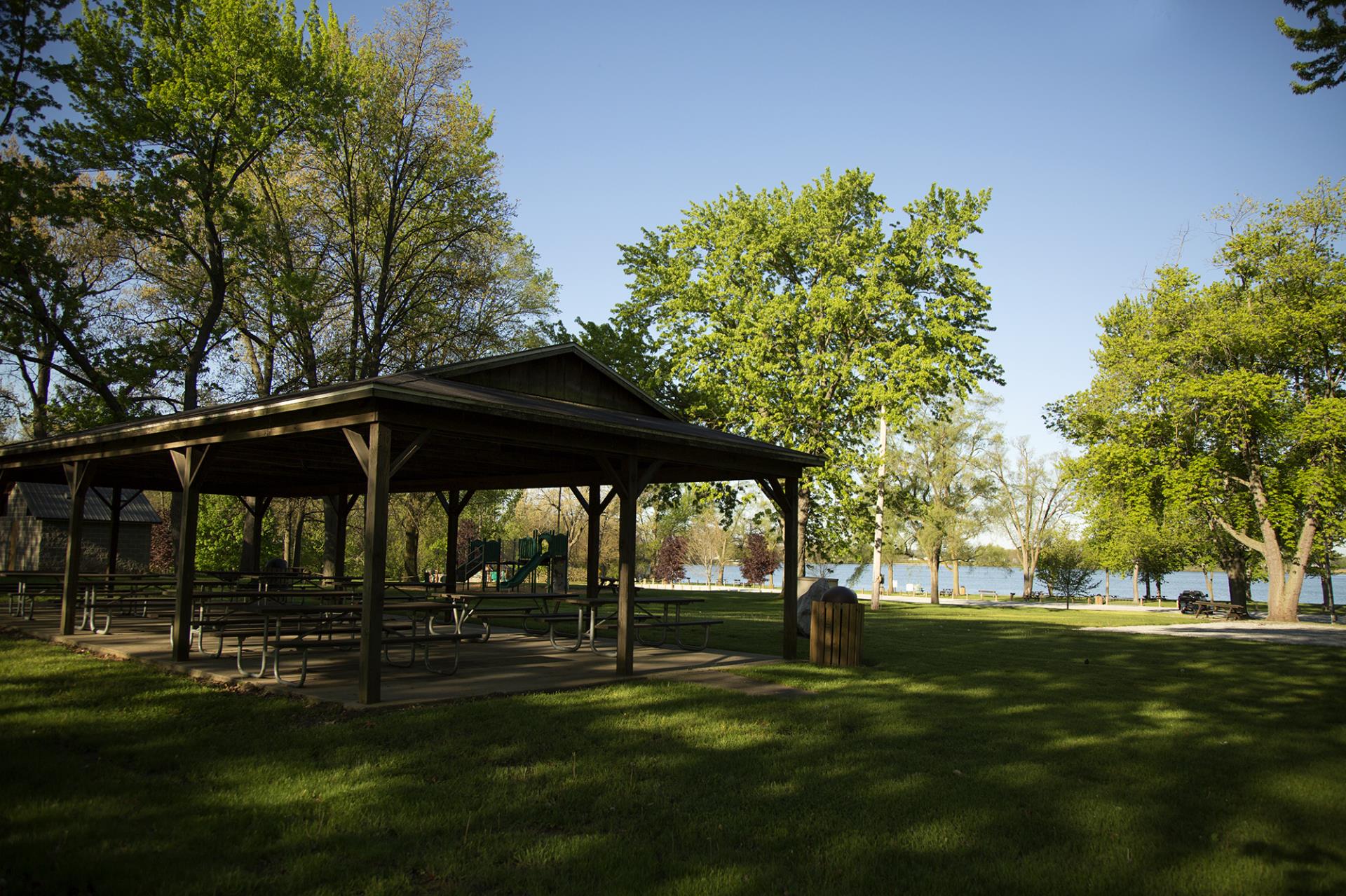 Large covered picnic shelter with attached tables in a lakeside park under clear skies.