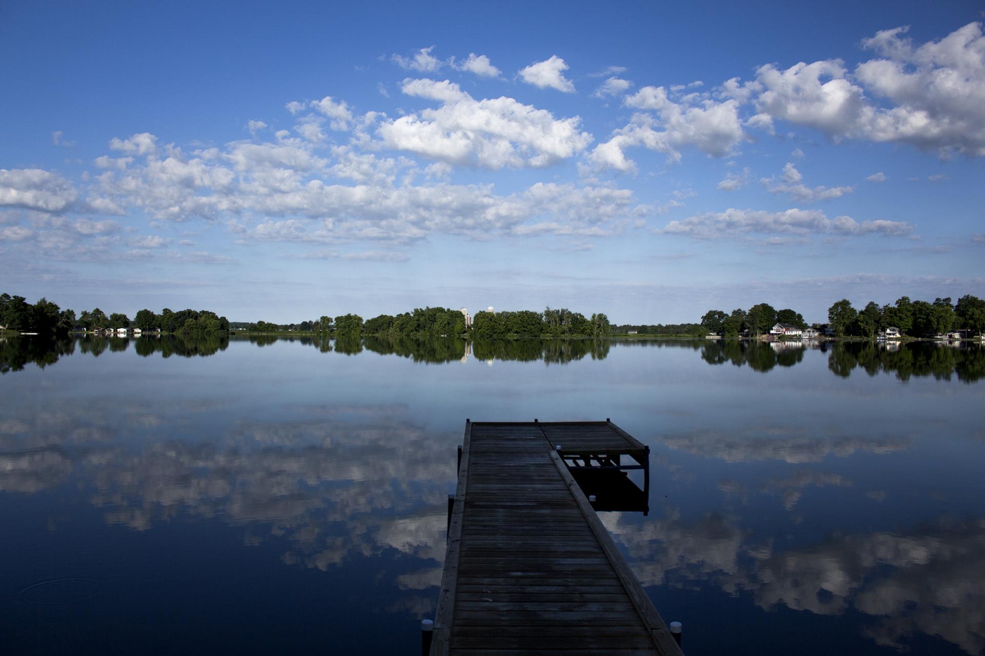 Wooden dock extending into a calm, mirror-like river with trees and houses lining the distant shoreline under a blue sky with white clouds.