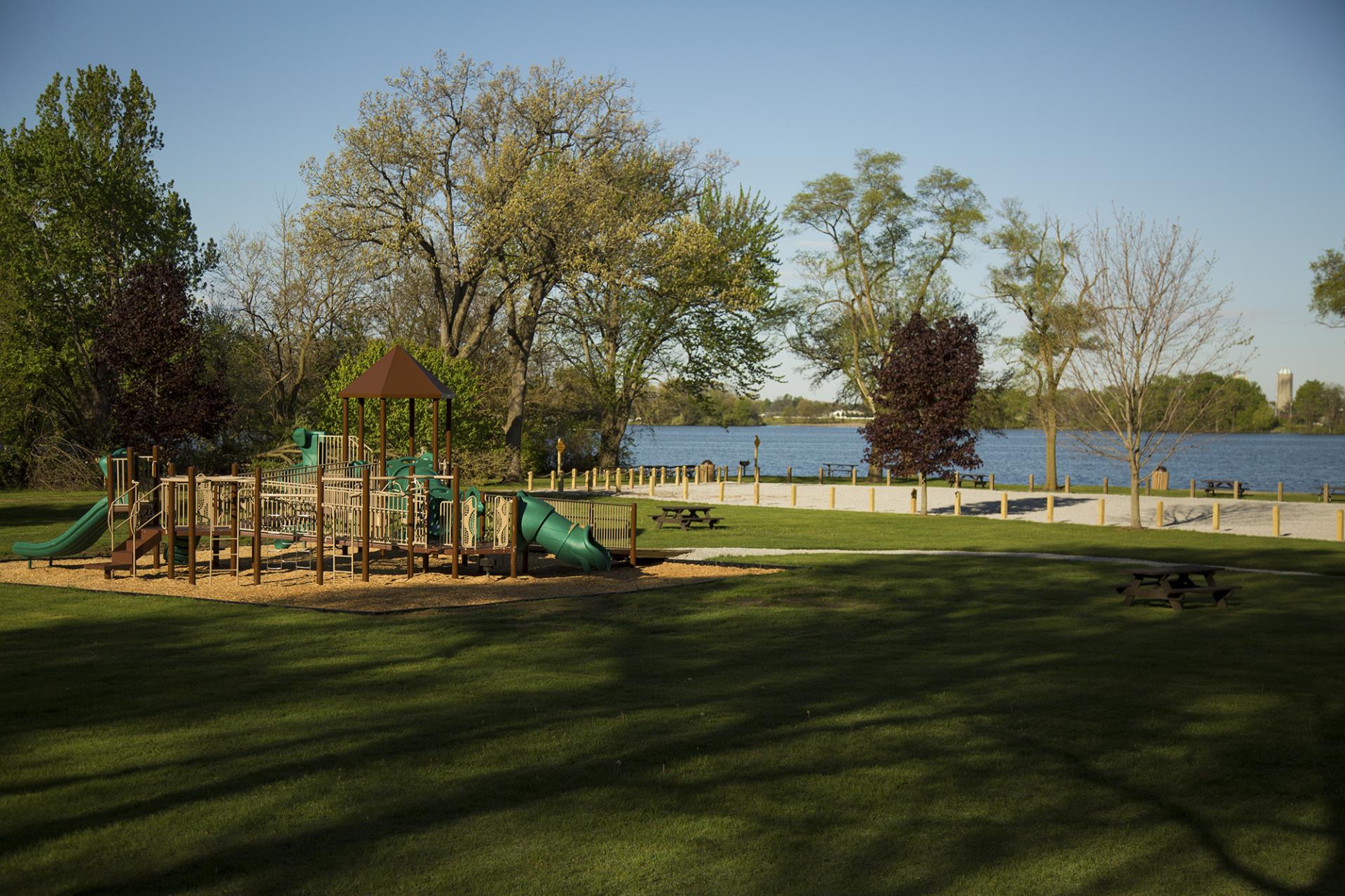 Waterfront park with playground, picnic tables, and grassy lawn bordered by trees on a clear day.