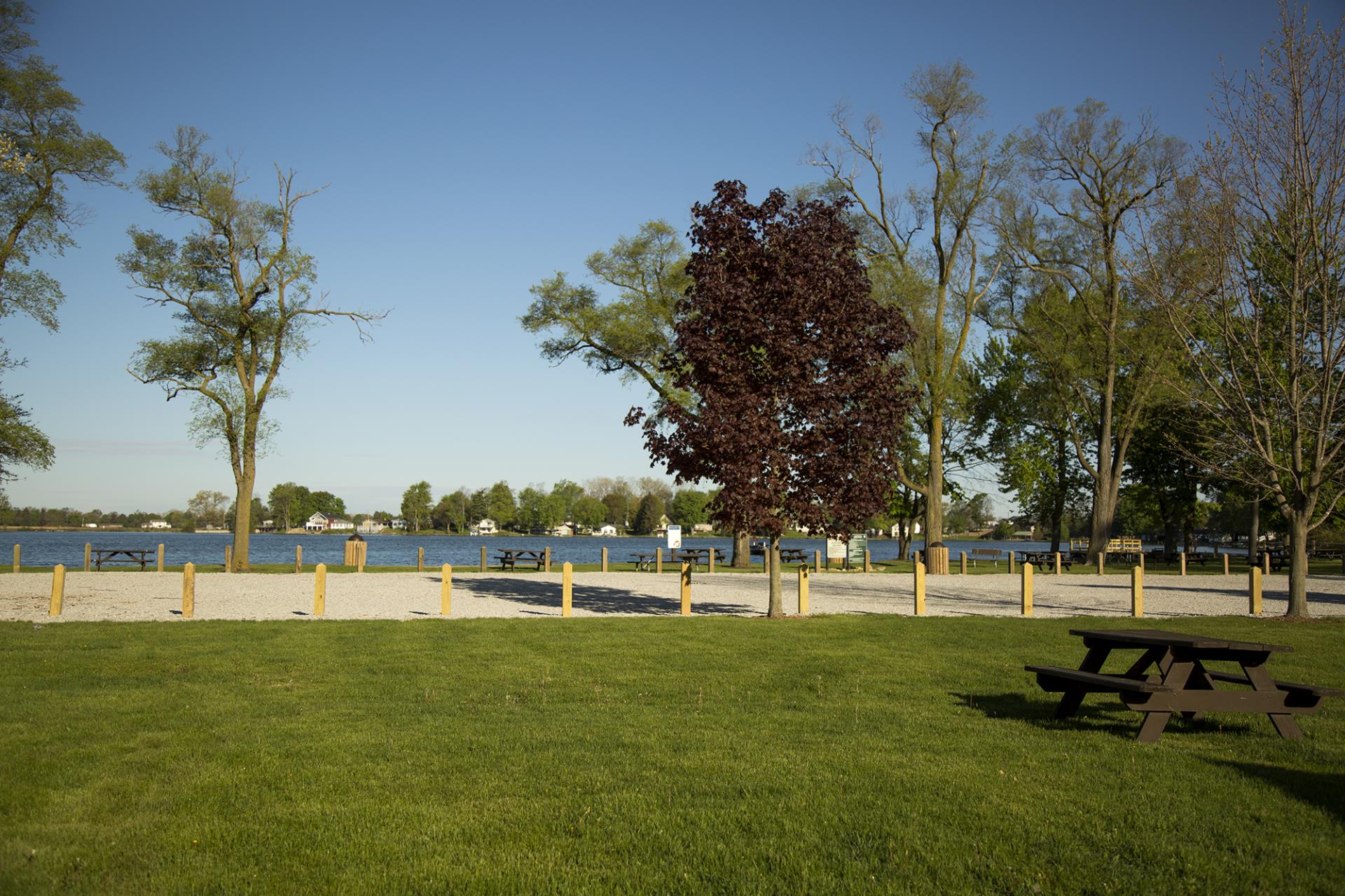 Waterfront park with sandy beach area, picnic tables, yellow bollards, trees, and residential homes across the water under clear sky.