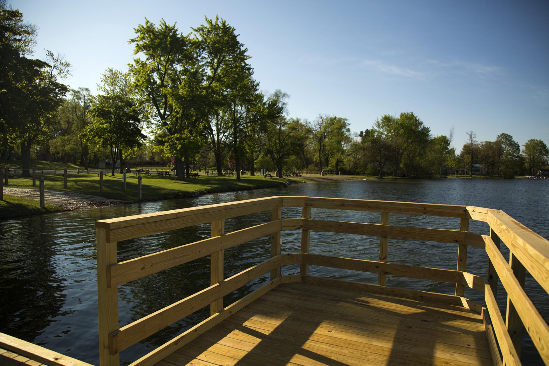 A wooden dock extends into a calm river with green trees and grassy park area visible along the shoreline.