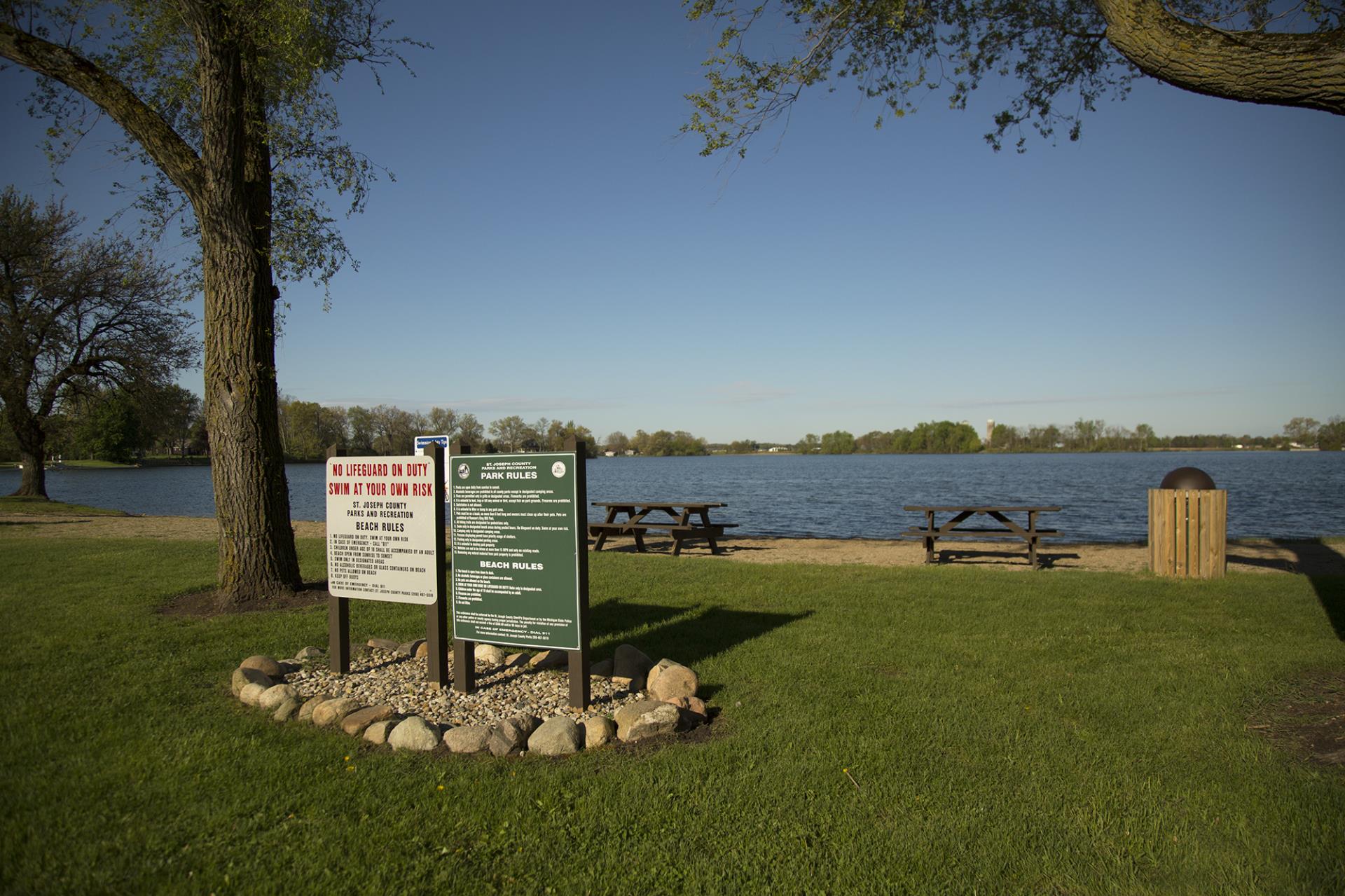 Waterfront park with informational signs, picnic tables, and a yellow trash receptacle along a grassy beach area.