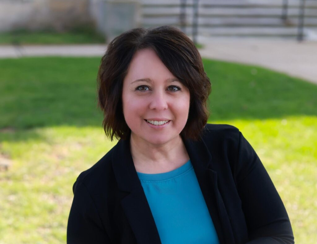 Woman with dark shoulder-length hair wearing a black blazer and blue shirt, smiling at camera with green lawn background.