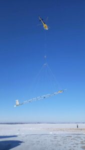 Yellow helicopter lowering a long beam structure over a frozen lake with personnel on the ice below.